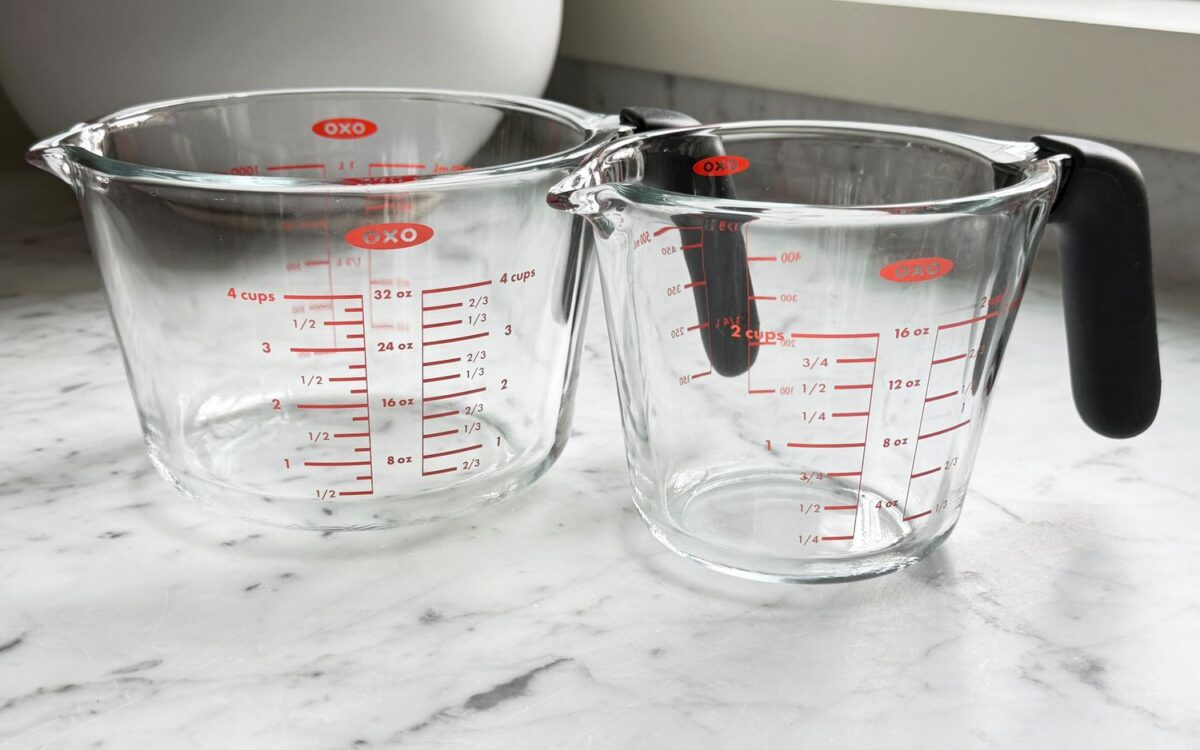 Clear measuring pitcher pouring water into a gallon jug on a kitchen counter, showing ounces to gallons